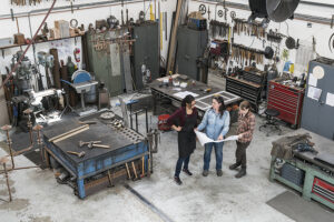 Vista de gran angular de tres mujeres de pie en un taller de metal, sosteniendo un plano técnico